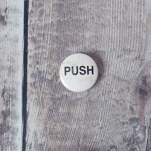 May include: A white, circular button with the word "PUSH" printed in black. The button is set against a weathered, grey wooden surface.