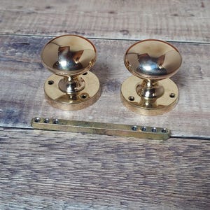 May include: A pair of polished brass door knobs and a brass bar with multiple holes. The knobs have a rounded top and a circular base. The items are displayed on a wooden surface.