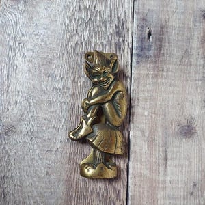 May include: A brass figurine of a seated goblin with a mischievous grin. The goblin is perched on a mushroom-shaped base. The figurine has a detailed design and a vintage aesthetic.