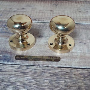 May include: Two polished brass door knobs with round, smooth tops and a circular base. The knobs are on a wooden surface.