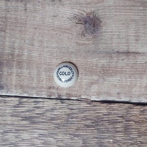 May include: A small, round, white ceramic bottle stopper with the word "COLD" in the center, surrounded by the text "YOUNG & MARTEN LTD. STRATFORD. FORDE". The stopper is set against a weathered, light brown wooden surface.