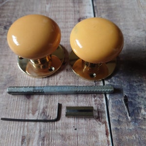 May include: Two brass door knobs with yellow ceramic round knobs. The knobs are shown with the mounting hardware, including a screw and an allen wrench.