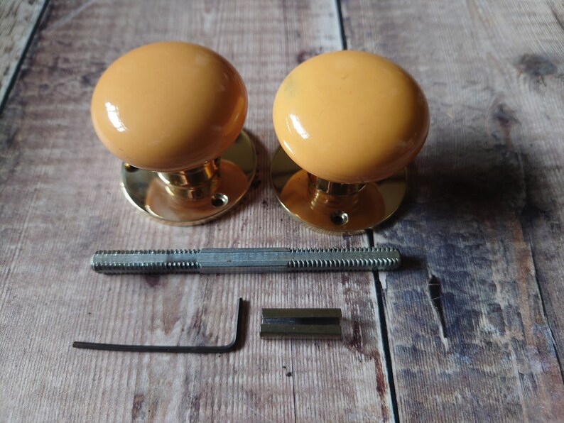 May include: Two brass door knobs with yellow ceramic round knobs. The knobs are shown with the mounting hardware, including a screw and an allen wrench.