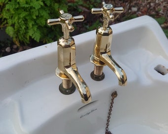 Puede incluir: Un lavabo de baño de porcelana blanca con dos grifos dorados y una cadena dorada unida a un tapón de desagüe. El lavabo tiene las palabras "Vintage Ware" impresas en la parte inferior.