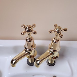 Könnte beinhalten: Zwei goldfarbene Wasserhähne mit kreuzförmigen Griffen sind an einem weißen Porzellanwaschbecken montiert. Das Waschbecken trägt die Aufschrift "Armitage Shanks" auf der Unterseite.