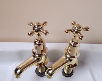 Puede incluir: Dos grifos de color dorado con manijas en forma de cruz están montados en un lavabo de porcelana blanca. El lavabo tiene las palabras "Armitage Shanks" impresas en la parte inferior.