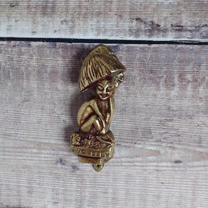 May include: A brass figurine of a seated figure under an umbrella. The figure is sitting on a base that reads "JOVELLY". The figurine has a gold finish and is set against a wooden background.