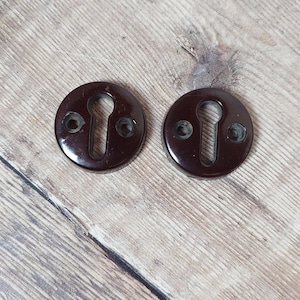 May include: Two dark brown, circular keyhole covers with a glossy finish. Each cover has a keyhole-shaped opening and two screw holes. The keyhole covers are set against a light brown, wood-grain background.