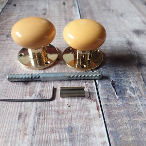 May include: Two brass door knobs with a yellow ceramic finish. The knobs are shown with mounting hardware, including a screw, an allen key, and a square-shaped piece of metal.