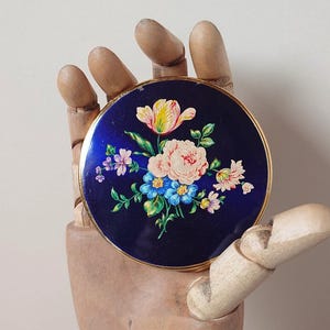 以下が含まれることがあります： 金色の縁取りが施された、ネイビーブルーの丸いコンパクト。ピンク、黄色、青の花柄が特徴です。木製の手に持たれており、ヴィンテージスタイルと装飾的な性質を示しています。