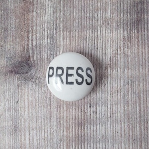 以下が含まれることがあります： 中央に太字の黒いサンセリフ体の大文字で「PRESS」という単語が印刷された、オフホワイトの丸いセラミック製ノブ。このオブジェクトは滑らかで光沢のある仕上げが施されており、光を反射するドーム型の凸面形状をしています。