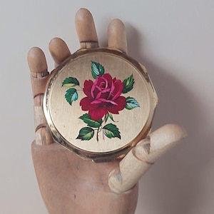 May include: A vintage gold-toned compact mirror with a floral design. The compact features a vibrant red rose with green leaves and blue accents. The compact is held by a wooden hand.