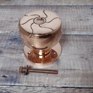 Peut inclure: Un bouton de porte en cuivre poli avec un motif tourbillonnant décoratif sur le dessus. Le bouton est monté sur une base circulaire, avec un boulon assorti à côté. La quincaillerie est présentée sur une surface en bois.