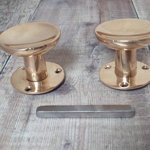 May include: Two antique brass door knobs with a matching metal bar handle. The knobs have a rounded top and a cylindrical base with screw holes. The metal bar is rectangular and silver in color. The items are on a wooden surface.