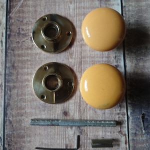 May include: A set of two yellow ceramic door knobs with brass backplates, a screw, an allen wrench, and a metal plate.