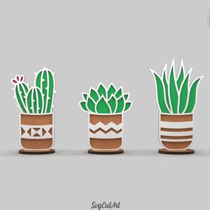 Puede incluir: Tres diseños de plantas suculentas cortadas con láser en macetas. Los diseños incluyen un cactus, una suculenta y una planta de aloe. Las plantas son verdes y las macetas son marrones con patrones geométricos blancos.