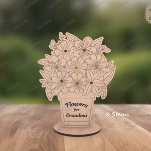 Könnte beinhalten: Holzblumenarrangement mit dem Text "Flowers for Grandma" und "8 Stems". Die Blumen befinden sich in einem topfförmigen Sockel, und das Design ist lasergeschnitten. Das Arrangement steht auf einem runden Sockel.