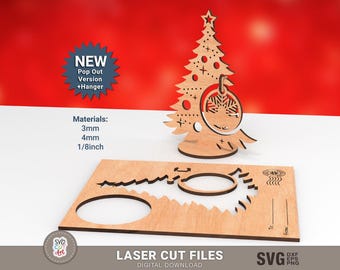 May include: A laser-cut wooden Christmas tree ornament with a star on top and a snowflake design. The ornament is displayed with a matching base and a separate piece with cut-out shapes. The text "NEW Pop Out Version + Hanger" is visible.