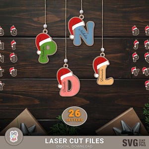 Peut inclure: Ornements de Noël en bois en forme de lettres, chacun orné d'un bonnet de Père Noël. Les lettres sont de différentes couleurs et sont suspendues par des ficelles. L'image comprend également des lettres individuelles et le texte "26 LETTERS".