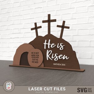 May include: A laser cut wooden Easter sign with three crosses and a tomb. The sign reads "He is Risen" with the scripture reference "Mathew 28:6". The tomb has a sign that reads "He is not here for He has risen just as He said."