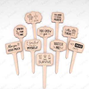 May include: Set of 11 wooden plant markers with funny and cute sayings. The markers feature a variety of designs, including a cactus, a succulent, and a heart. The sayings include "I need a drink", "You grow girl", "Aloe you vera much", "I gorled myself", "Water me!" and "Not dead (yet)".
