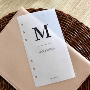 Puede incluir: Una página de planificador blanca translúcida con una letra negra "M" y el texto "MAY JORDAN" y "MAY PAPER CO."