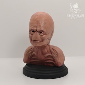 May include: A tan-colored humanoid bust sculpture featuring a smooth, hairless head, deep-set eyes, and prominent facial wrinkles. The figure has a defined neck and shoulder structure, resting on a circular black base. The text SHADOWREALM STUDIO is visible.