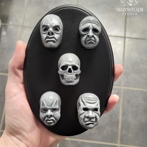Könnte beinhalten: Eine ovale schwarze Plakette mit fünf kleinen, grauen, skulptierten Gesichtern in einem Muster. Das Mittelstück ist ein menschlicher Schädel, umgeben von vier verschiedenen, ausdrucksstarken Gesichtern. Der Text SHADOWREALM STUDIO ist weiß gedruckt.