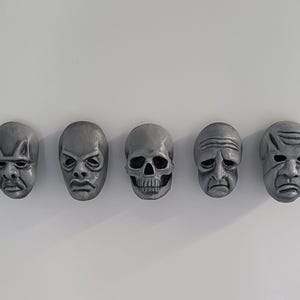 Könnte beinhalten: Fünf graue, geformte Gesichtsmasken mit unterschiedlichen Ausdrücken sind horizontal vor einem weißen Hintergrund angeordnet. Die Masken umfassen einen Schädel, ein dämonisches Gesicht und Gesichter mit gerunzelten Augenbrauen und Grimassen. Die Masken sind wahrscheinlich dekorativ.