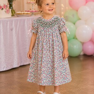 May include: A young child wearing a floral print dress with a green embroidered neckline and short sleeves tied with pink ribbons. The dress is paired with white Mary Jane shoes. A pink bow adorns the child's hair.
