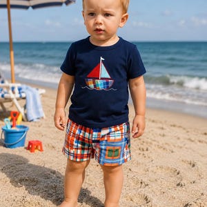 May include: A young child wearing a navy blue t-shirt with a sailboat design and plaid shorts stands on a sandy beach. The sailboat has a red sail and a colorful plaid body. The shorts are orange, blue, and white plaid.