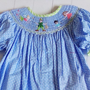 Puede incluir: Vestido azul claro con lunares blancos, mangas cortas abullonadas y cuello redondo. El cuello y los pu&ntilde;os de las mangas est&aacute;n ribeteados en verde. El corpi&ntilde;o presenta un dise&ntilde;o bordado de Peter Pan y otros personajes.