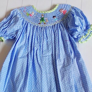 Puede incluir: Vestido azul de lunares con bordados de personajes de Peter Pan alrededor del cuello. El vestido presenta mangas cortas abullonadas con ribete verde y un escote con ribete verde. La tela es azul claro con lunares blancos.