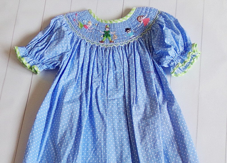 Puede incluir: Vestido azul de lunares con bordados de personajes de Peter Pan alrededor del cuello. El vestido presenta mangas cortas abullonadas con ribete verde y un escote con ribete verde. La tela es azul claro con lunares blancos.