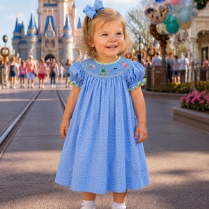 Puede incluir: Una ni&ntilde;a peque&ntilde;a con un vestido azul claro de lunares con corpi&ntilde;o smock y detalles bordados. El vestido tiene mangas cortas con ribete verde y se combina con zapatos blancos y un lazo azul. El fondo muestra un castillo y globos.
