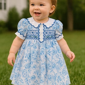 May include: A smiling toddler wearing a light blue dress with a white collar and a floral rose pattern. The dress has short sleeves and a smocked bodice. A large pink bow adorns the child's head. The child is standing on green grass.