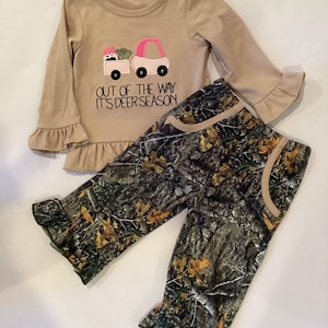 May include: A tan long-sleeved shirt with a pink and white truck graphic and the text "OUT OF THE WAY IT'S DEER SEASON." The shirt has ruffle sleeves and hem. The set includes camouflage print pants.