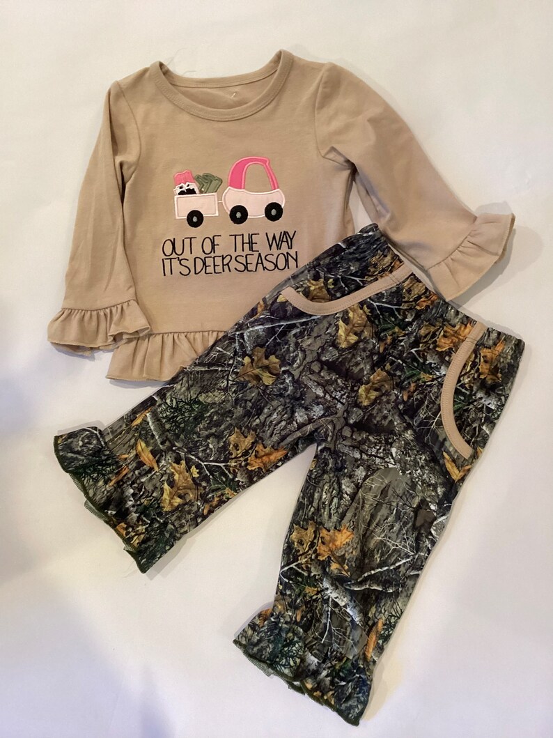 May include: A tan long-sleeved shirt with a pink and white truck graphic and the text "OUT OF THE WAY IT'S DEER SEASON." The shirt has ruffle sleeves and hem. The set includes camouflage print pants.