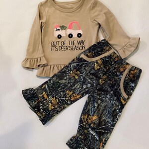 May include: Tan long-sleeved shirt and camouflage pants set. The shirt features a pink and white truck graphic with the text "OUT OF THE WAY IT'S DEER SEASON". The pants have a camouflage pattern with tan accents.