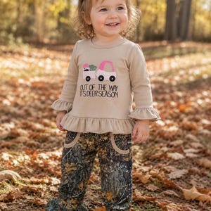 May include: A tan long-sleeved shirt with a pink truck graphic and the text "OUT OF THE WAY IT'S DEER SEASON." The shirt has ruffled sleeves and is paired with camouflage pants with ruffled bottoms. A green bow is in the hair.