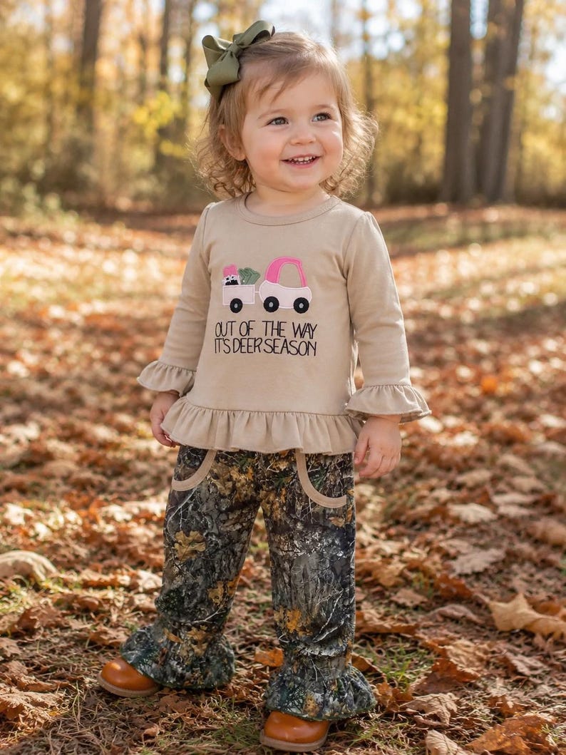 May include: A tan long-sleeved shirt with a pink truck graphic and the text "OUT OF THE WAY IT'S DEER SEASON." The shirt has ruffled sleeves and is paired with camouflage pants with ruffled bottoms. A green bow is in the hair.