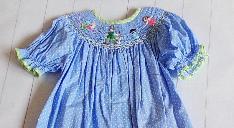 Puede incluir: Vestido azul claro con lunares blancos, mangas cortas abullonadas y cuello redondo. El cuello y los pu&ntilde;os de las mangas est&aacute;n ribeteados en verde. El corpi&ntilde;o presenta personajes bordados, incluyendo Peter Pan, Campanilla y otros.