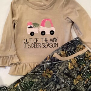 May include: A tan long-sleeved shirt with a pink and white truck graphic and the text "OUT OF THE WAY IT'S DEER SEASON." The shirt has ruffle sleeves and is paired with camouflage print pants.
