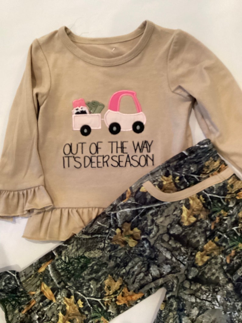May include: A tan long-sleeved shirt with a pink and white truck graphic and the text "OUT OF THE WAY IT'S DEER SEASON." The shirt has ruffle sleeves and is paired with camouflage print pants.