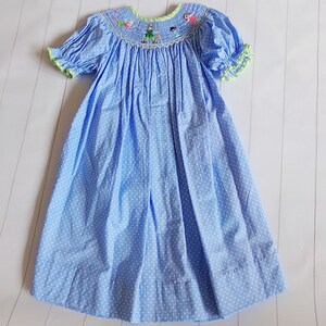 Puede incluir: Vestido azul claro con lunares blancos, mangas cortas abullonadas y cuello redondo. El cuello y los pu&ntilde;os tienen un ribete verde. El corpi&ntilde;o presenta personajes bordados. El vestido es de tejido ligero.