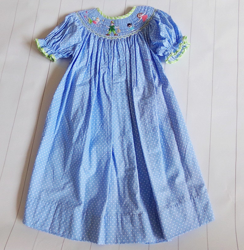 Puede incluir: Vestido azul claro con lunares blancos, mangas cortas abullonadas y cuello redondo. El cuello y los pu&ntilde;os tienen un ribete verde. El corpi&ntilde;o presenta personajes bordados. El vestido es de tejido ligero.