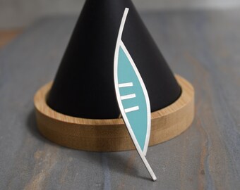 May include: A silver leaf-shaped brooch with a teal blue enamel inlay and three white lines. The brooch is sitting on a wooden base.