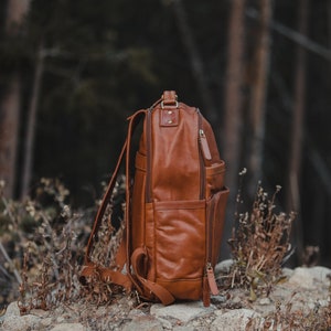 May include: A brown leather backpack with multiple compartments and zippers. The bag has adjustable shoulder straps and a top handle. It is sitting on a rocky surface with a blurred forest background.