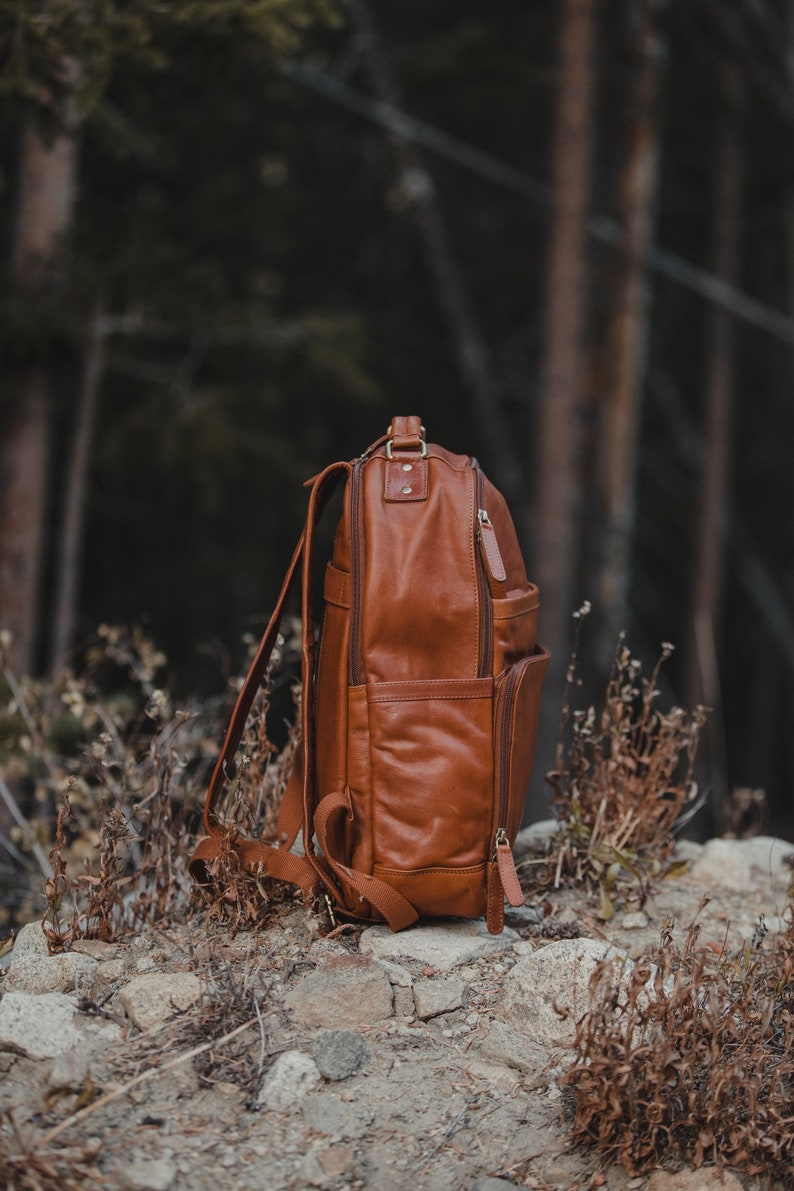 May include: A brown leather backpack with multiple compartments and zippers. The bag has adjustable shoulder straps and a top handle. It is sitting on a rocky surface with a blurred forest background.
