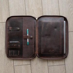 May include: A dark brown leather portfolio case featuring a full-length zipper, multiple interior card slots, a pen holder with a white and teal pen, and a dedicated sleeve containing a laptop. A small bear logo is embossed on the interior.